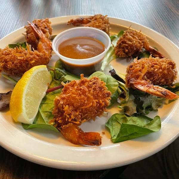 Six crispy coconut shrimp with dipping sauce, a lemon wedge, and greens on a white plate.